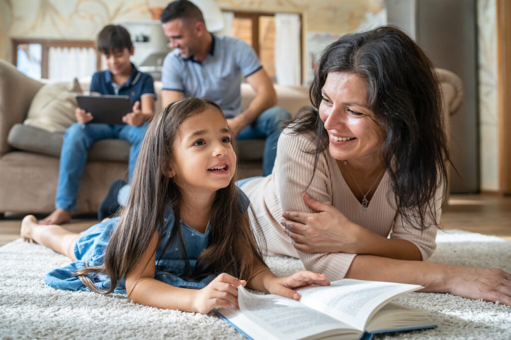 Mum Is Reading A Book On The Floor With Her Daughter, While Dad And Son Are Sitting On The Sofa